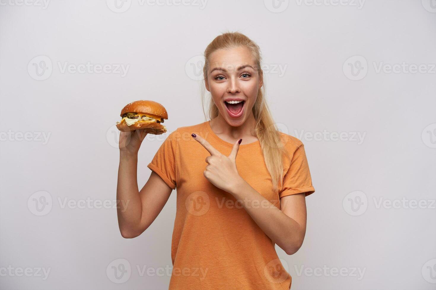 felicissimo bellissimo giovane bionda femmina con coda di cavallo acconciatura guardare a telecamera con largo occhi e bocca ha aperto e mostrando su gustoso Hamburger nel sua mano, isolato al di sopra di bianca sfondo foto