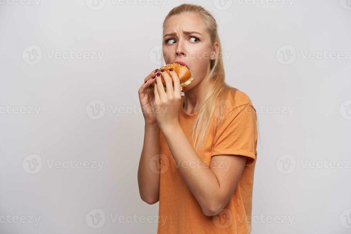 ritratto di confuso giovane bella bionda femmina con casuale acconciatura guardare in modo emozionante a parte e arrotondamento occhi mentre avendo pranzo con Fast food, isolato al di sopra di bianca sfondo foto