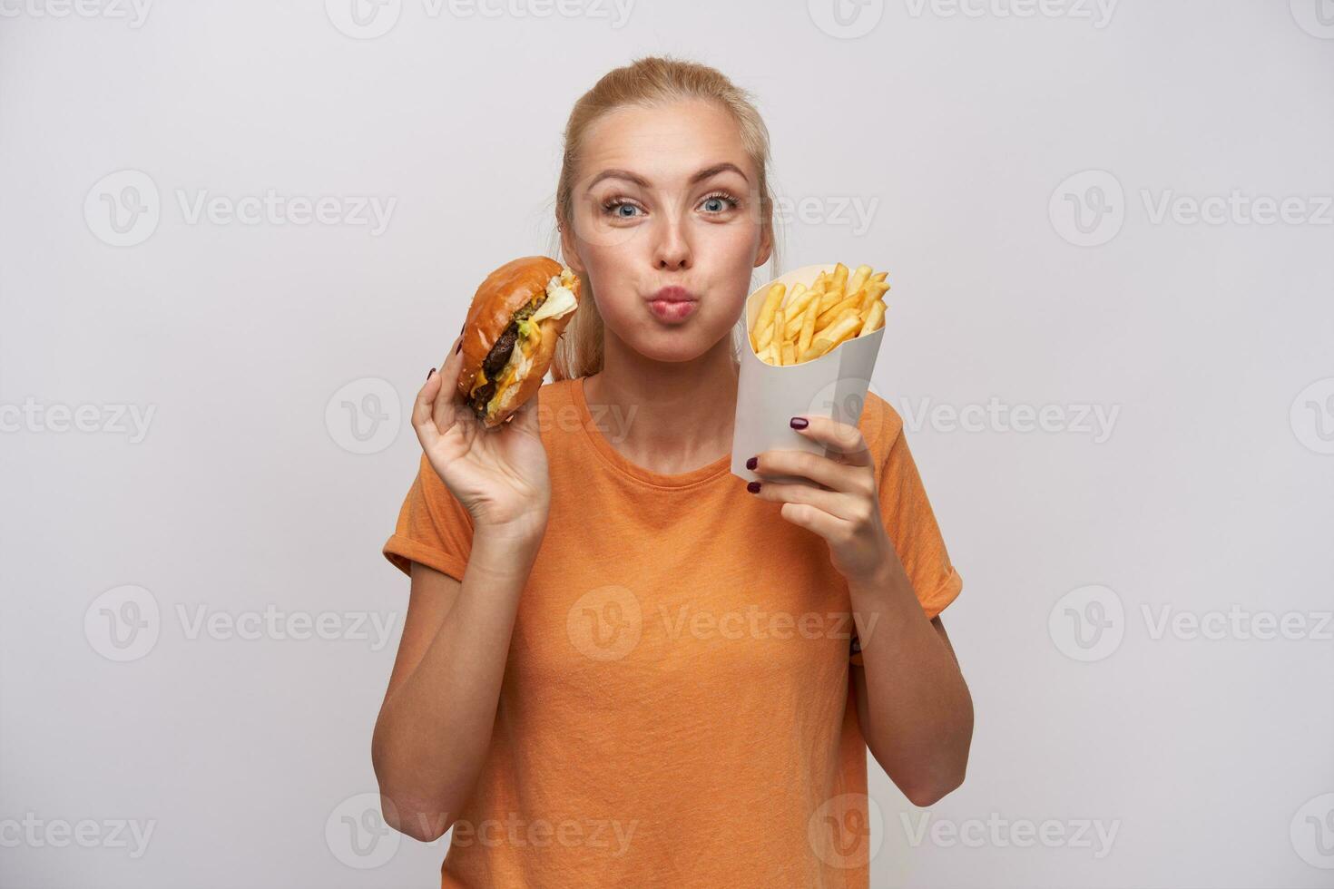 ritratto di affascinante bella giovane bionda donna Tenere Rifiuto cibo e guardare con gioia per telecamera, sbuffando su guance e essere eccitato di gustoso cena, isolato al di sopra di bianca sfondo foto