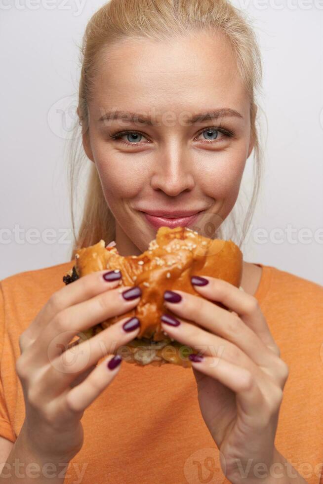 avvicinamento di bellissimo occhi azzurri giovane bionda donna mangiare veloce cibo e guardare allegramente a telecamera, sorridente piacevolmente mentre in posa contro bianca sfondo nel casuale Abiti foto