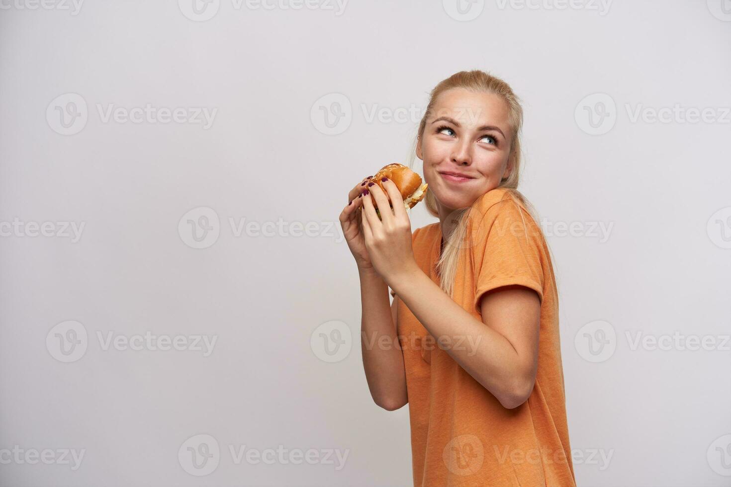 studio tiro di Affamato lieta giovane bionda femmina nel arancia maglietta guardare positivamente a parte con bocca pieno di cibo, Tenere fresco hamburger mentre in piedi al di sopra di bianca sfondo foto