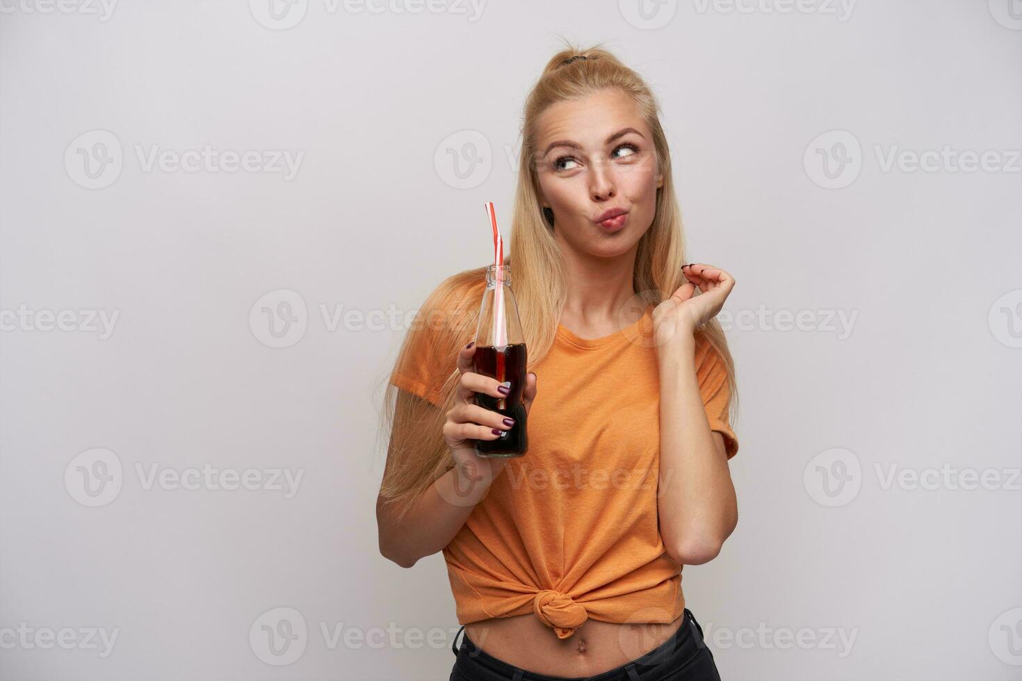 studio foto di positivo attraente giovane bionda signora con coda di cavallo acconciatura inseguendo labbra mentre guardare a parte con bottiglia di bibita nel sollevato mano, isolato al di sopra di bianca sfondo