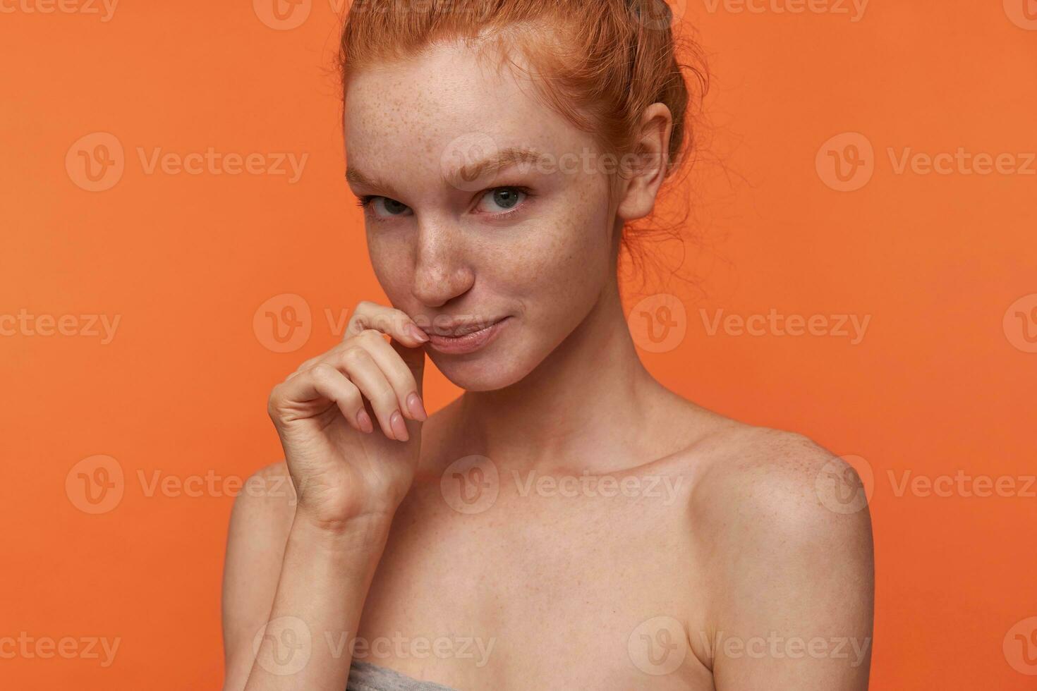 avvicinamento di positivo timido giovane lettore donna con casuale acconciatura guardare a telecamera delicatamente e conservazione mano su guancia, in piedi al di sopra di arancia sfondo con piegato labbra foto