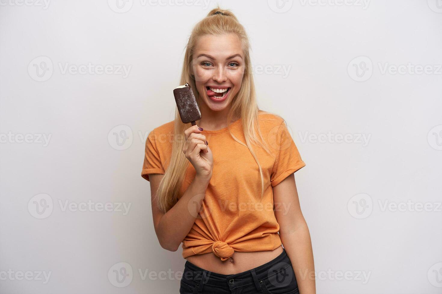 divertente tiro di gioioso bella giovane lungo dai capelli bionda donna nel arancia maglietta mostrando felicemente lingua per telecamera mentre in posa al di sopra di bianca sfondo con gelato foto