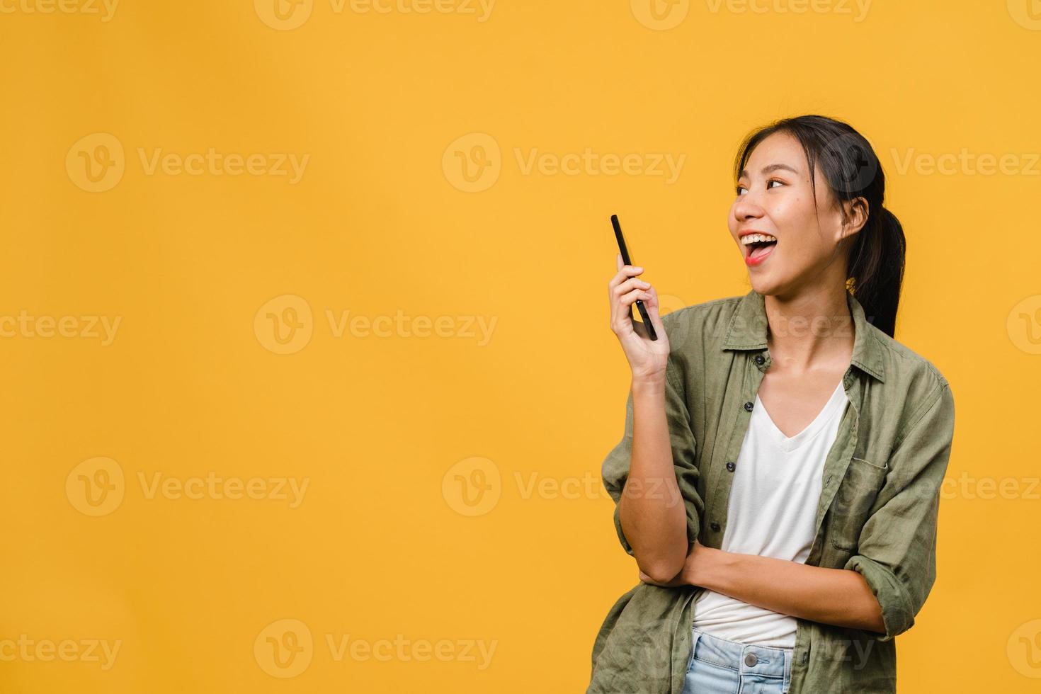 giovane donna asiatica che usa il telefono con un'espressione positiva, sorride ampiamente, vestita con abiti casual sentendosi felici e in piedi isolato su sfondo giallo. felice adorabile donna felice esulta successo. foto