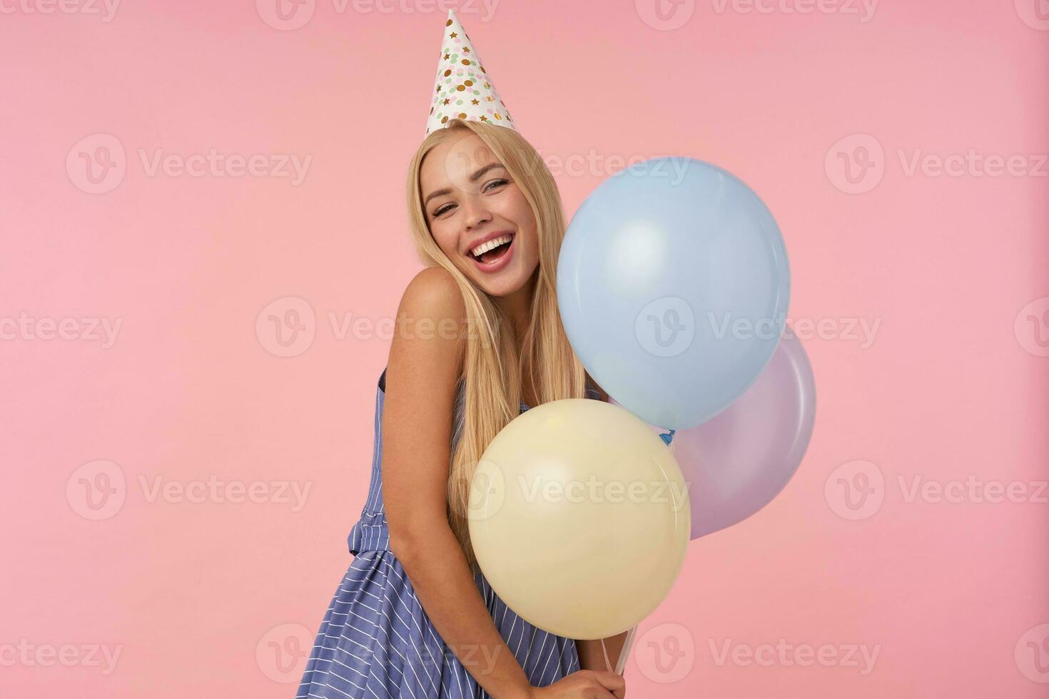 Contentissimo giovane bionda donna gioia mentre in posa nel multicolore aria palloncini, indossare festivo Abiti e cono cappello, isolato al di sopra di rosa sfondo, mostrando sua piacevole emozioni foto