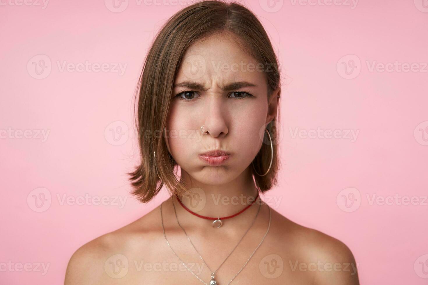 avvicinamento di giovane grave guardare corto dai capelli brunetta donna sbuffando su sua guance e accigliato sopracciglia mentre guardare a telecamera, isolato al di sopra di rosa sfondo foto