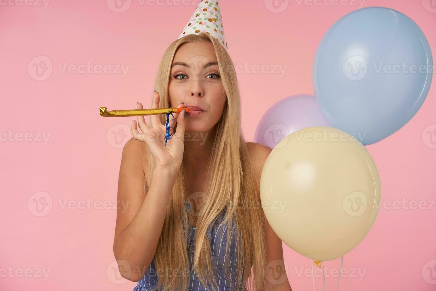 gioioso lungo dai capelli bionda donna nel blu estate vestito e compleanno berretto Tenere mazzo di elio palloncini, guardare a telecamera allegramente e soffiaggio festa corno, isolato al di sopra di rosa sfondo foto