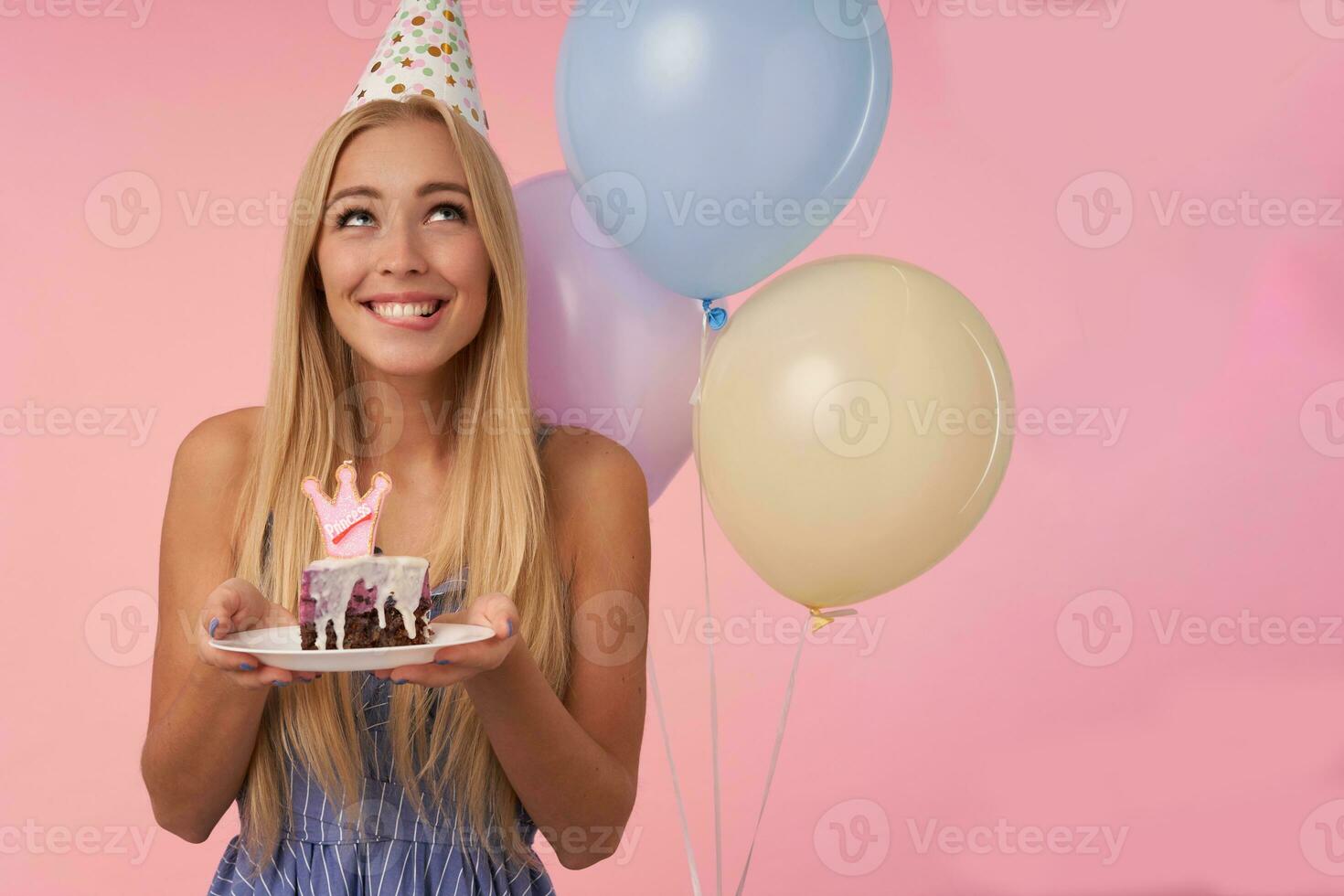 sognante bella lungo dai capelli bionda femmina conservazione pezzo di torta con candela e sorridente felicemente, in posa al di sopra di rosa sfondo con mazzo di multicolore elio palloncini foto