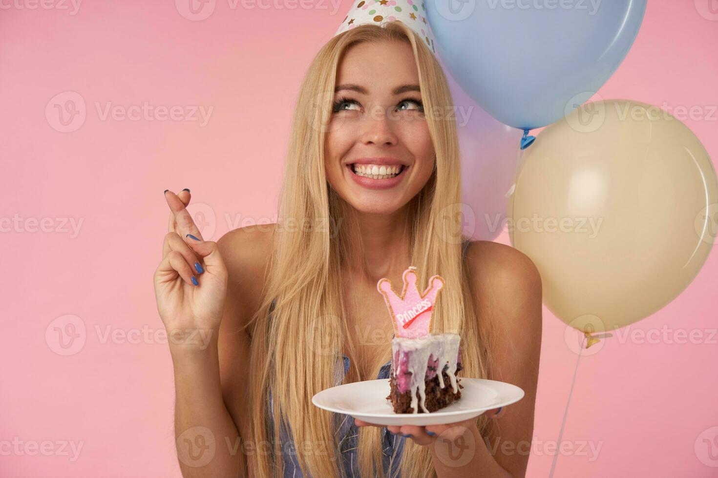 allegro giovane lungo dai capelli femmina con lungo bionda capelli attraversamento dita mentre fabbricazione desiderio su sua compleanno, Tenere pezzo di torta con candela al di sopra di multicolore aria palloncini e rosa sfondo foto