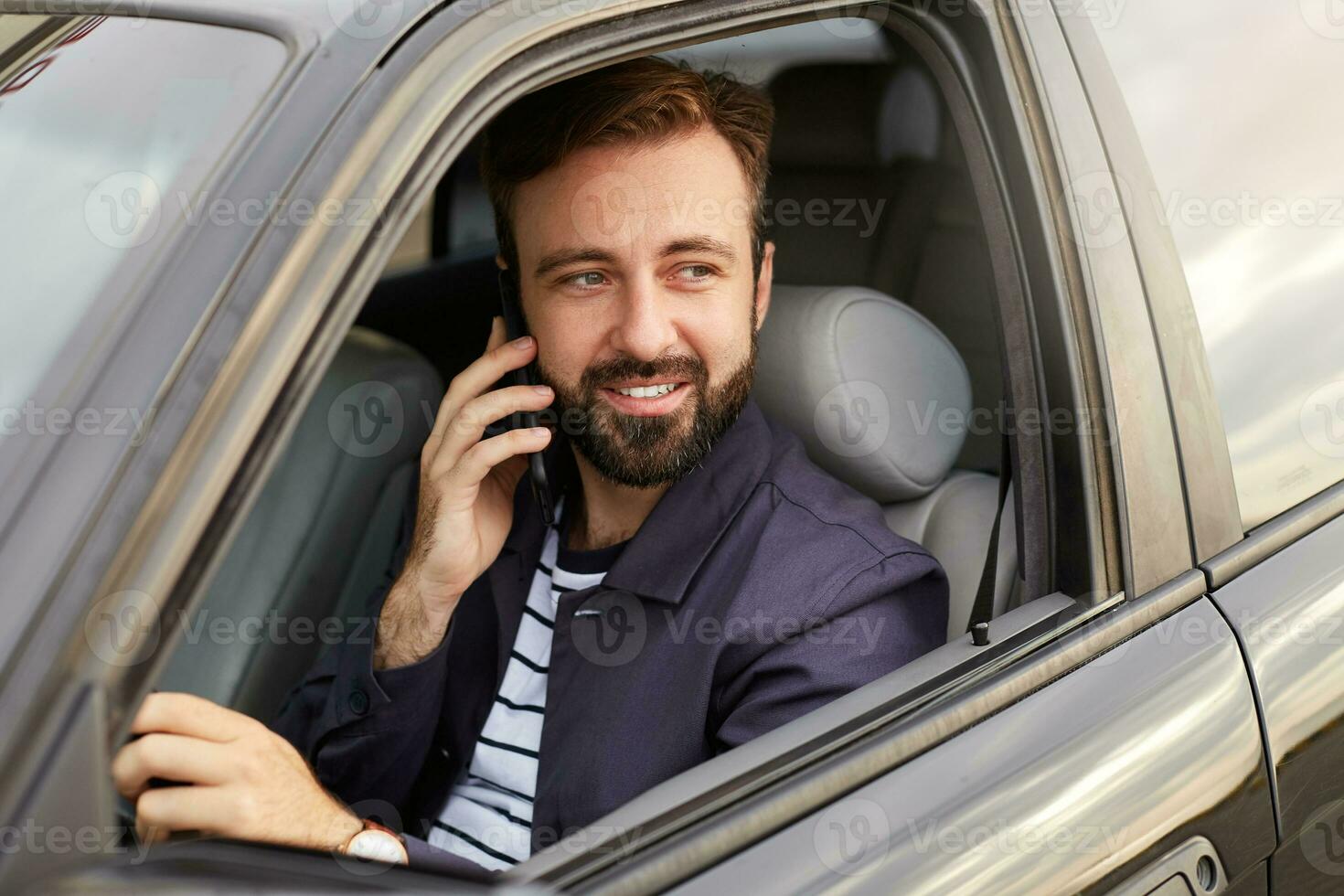 foto di di giovane attraente riuscito barbuto uomo nel un' blu giacca e a strisce maglietta, si siede dietro a il ruota di il auto e aspetta per un' risposta su il telefono.