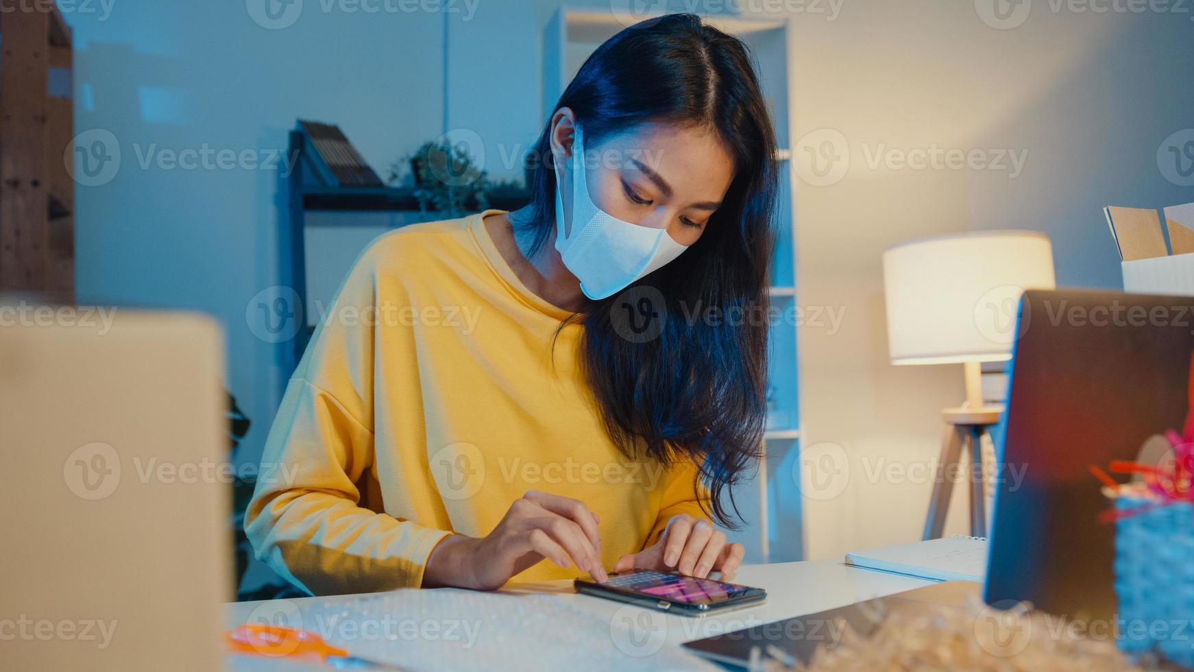 la giovane donna d'affari asiatica indossa la maschera per il viso controlla l'ordine di acquisto sullo smartphone e lo stock nel computer in ufficio a casa durante la notte. piccolo imprenditore, consegna al mercato online, concetto di lifestyle freelance. foto