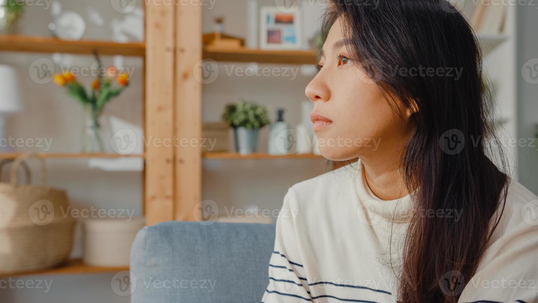 premurosa signora asiatica si siede abbracciando le ginocchia al divano nel soggiorno a casa guarda fuori con sentirsi solo, triste adolescente depresso trascorrere del tempo da solo stare a casa, distanza sociale, quarantena del coronavirus. foto