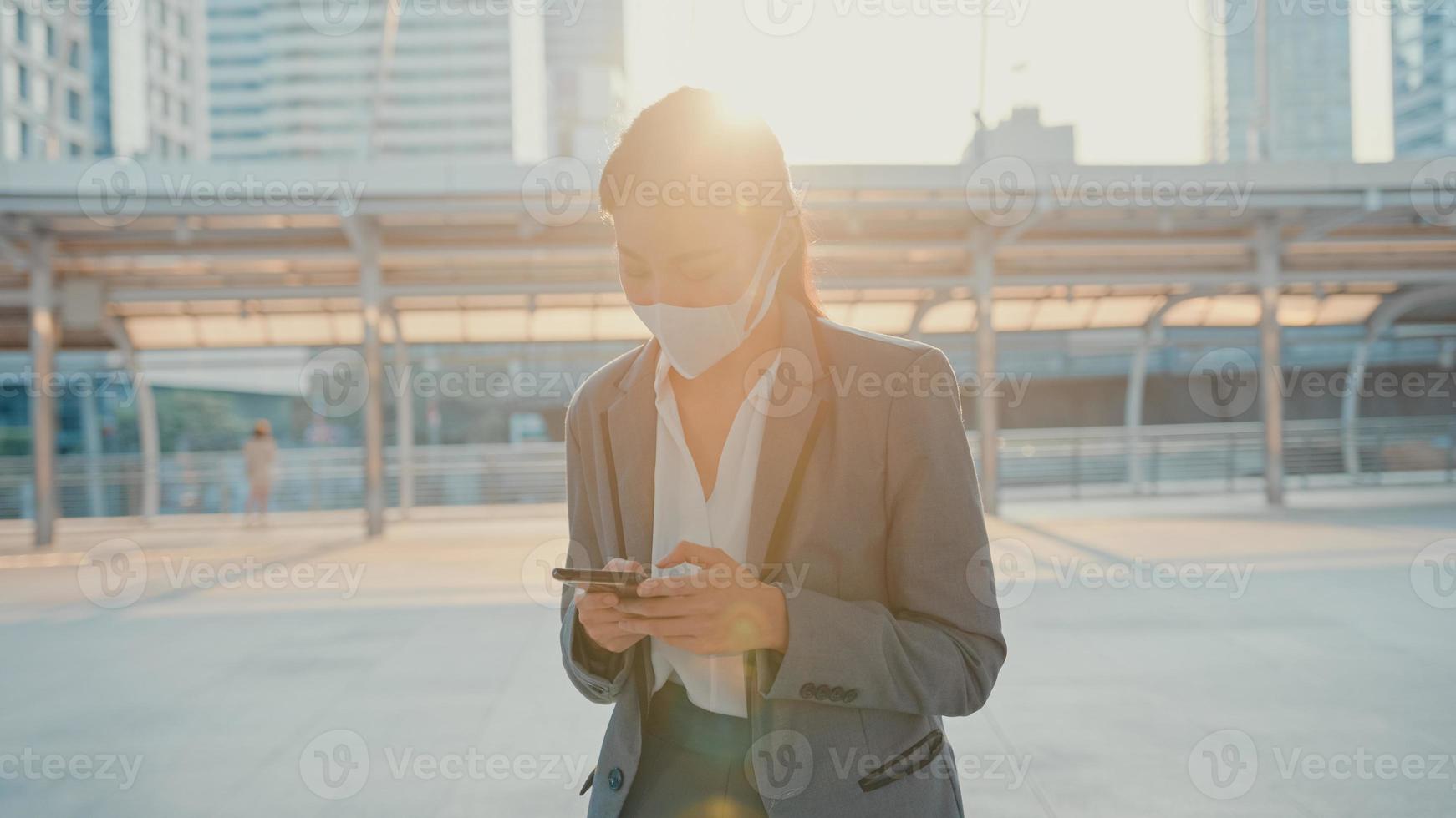 la giovane donna d'affari asiatica in abiti da ufficio alla moda indossa una maschera medica usando il telefono mentre cammina da sola all'aperto nella città urbana. affari in movimento, distanza sociale per prevenire la diffusione del concetto di covid-19. foto