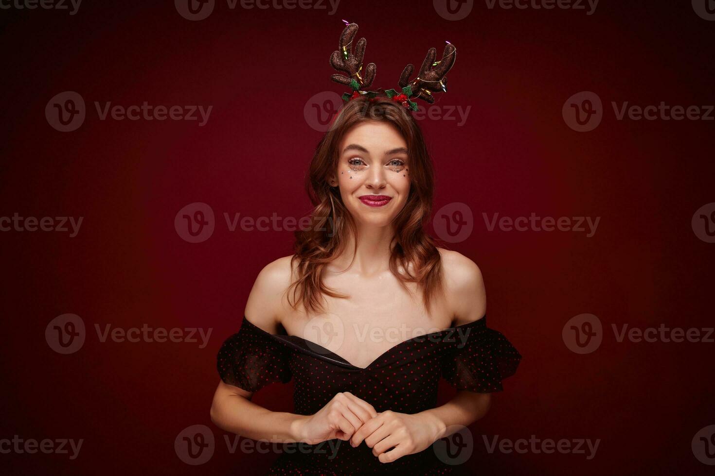 positivo bellissimo giovane Marrone dai capelli donna indossare elegante nero vestito con rosso puntini mentre in piedi al di sopra di chiaretto sfondo, gioia simpatico nuovo anno festa insieme con amici foto