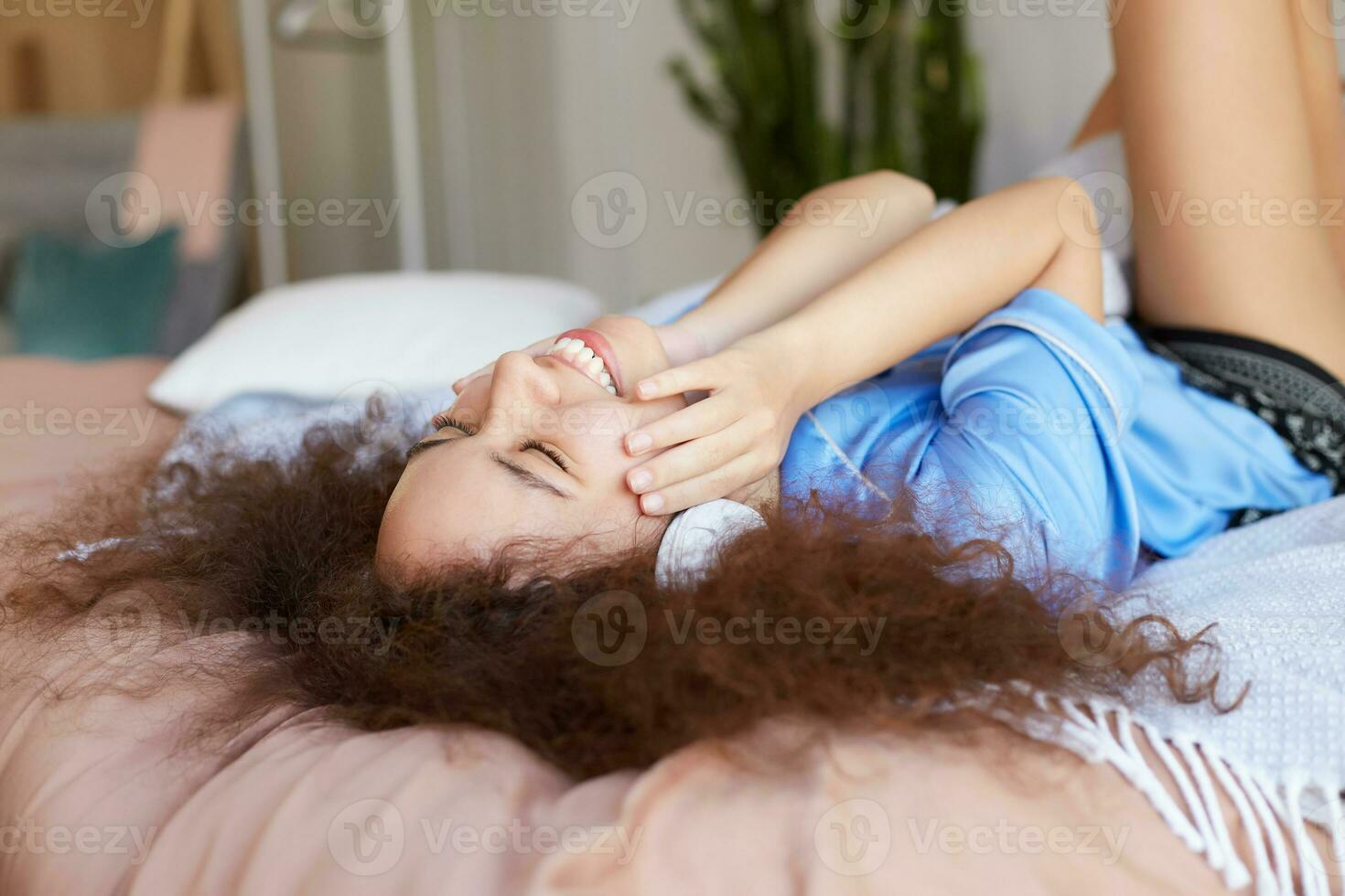 gioioso Riccio mulatto ragazza dire bugie su il letto con sua testa giù e chiuso occhi, ascoltando preferito canzone nel cuffia, a grandi linee sorridente e tocchi guance, sembra contento. foto