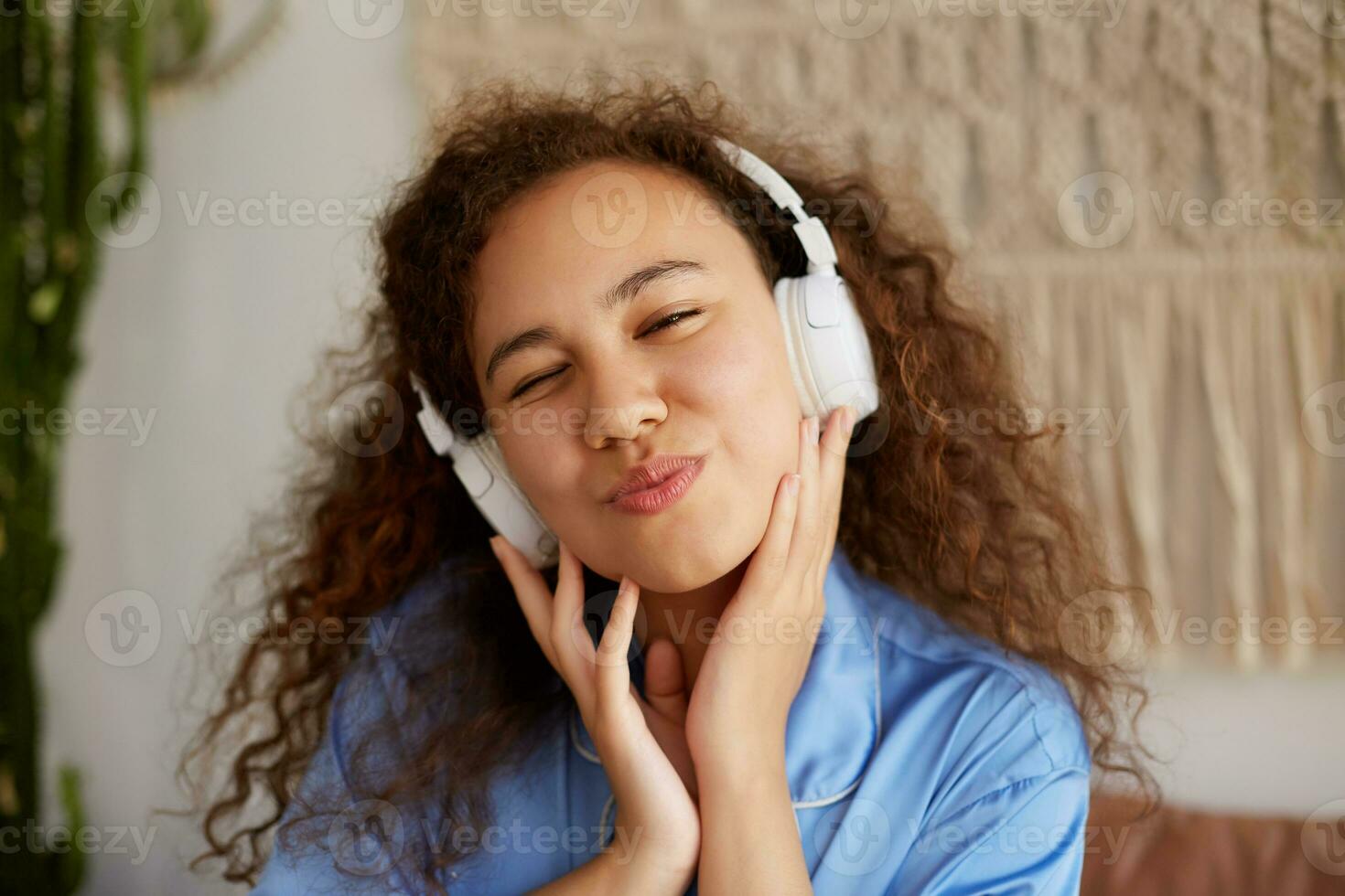 contento Riccio mulatto giovane signora ubicazione su il letto, occhiolino e ascoltando preferito musica nel cuffia, godendo il domeniche mattina. foto