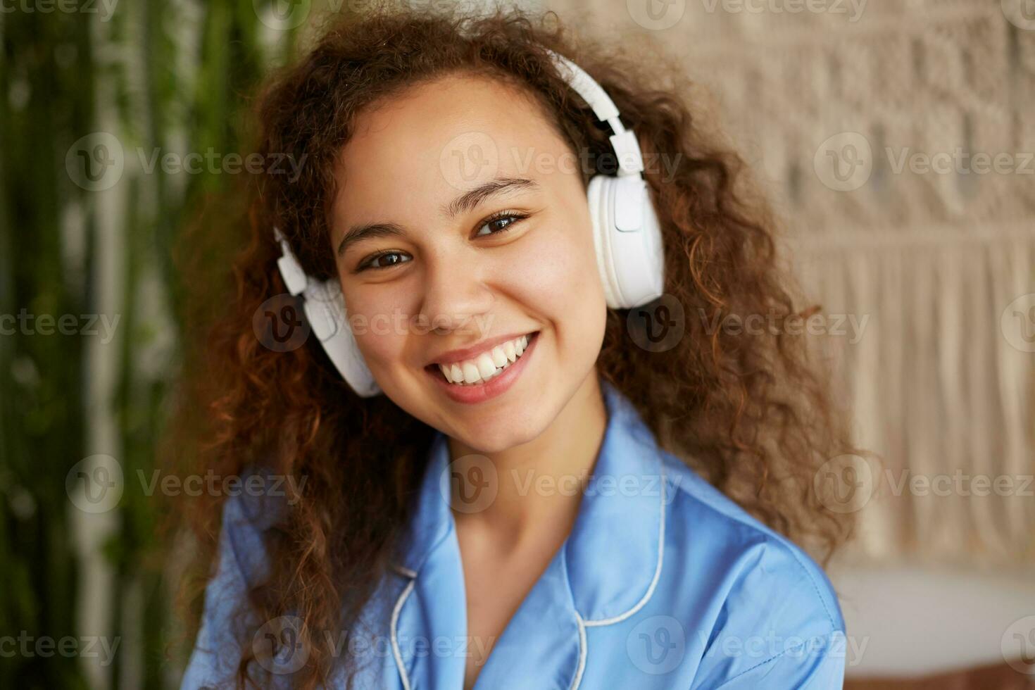 vicino su di contento Riccio mulatto giovane donna, a grandi linee sorrisi e ascoltando preferito canzone nel cuffia, godendo il musica e domeniche mattina. foto