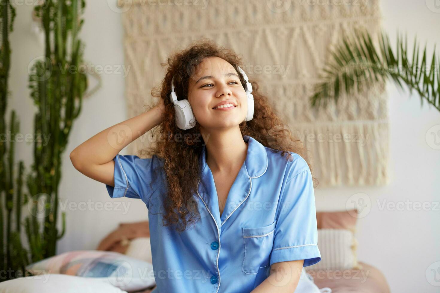 ritratto di Riccio giovane simpatico mulatto signora, a grandi linee sorrisi con chiuso occhi, ascoltando preferito musica nel cuffia, godendo il domeniche mattina, sembra contento e allegro. foto