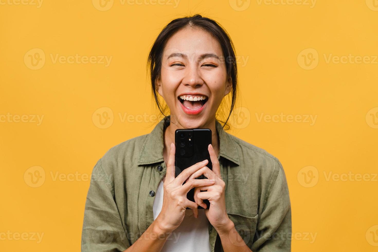 sorpresa giovane signora asiatica che utilizza il telefono cellulare con espressione positiva, sorride ampiamente, vestita con abiti casual e guardando la telecamera su sfondo giallo. felice adorabile donna felice esulta successo. foto