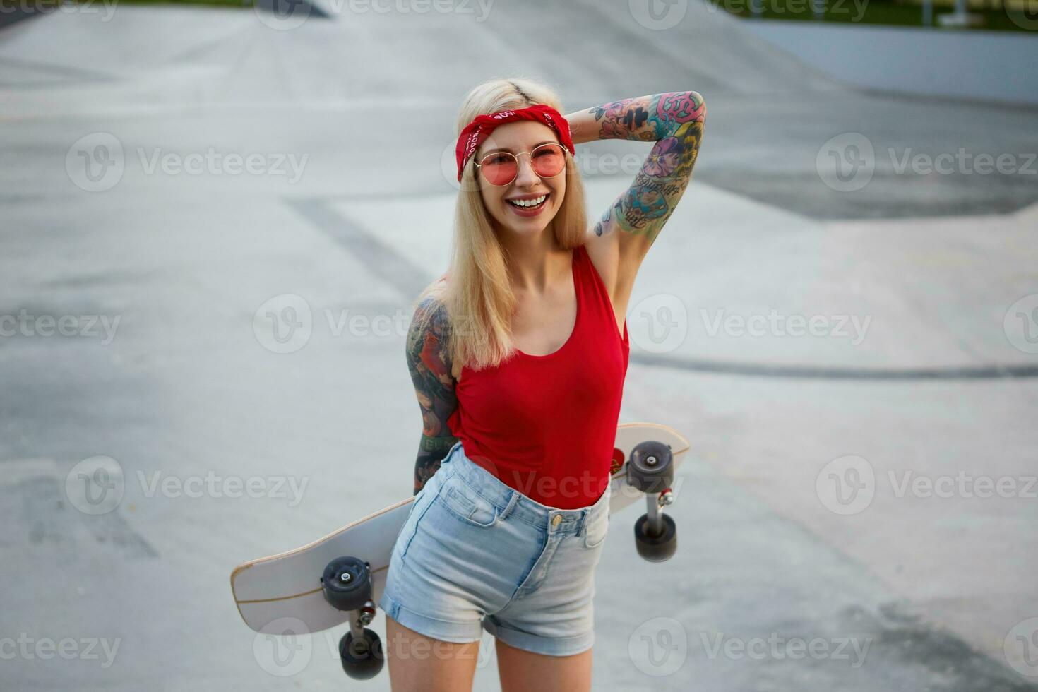 simpatico bionda signora con tatuato braccia vestito nel un' rosso maglietta e denim corti, con un' a maglia bandana su sua testa, nel rosso occhiali, Tenere un' longboard nel sua mani e a grandi linee sorridente. foto