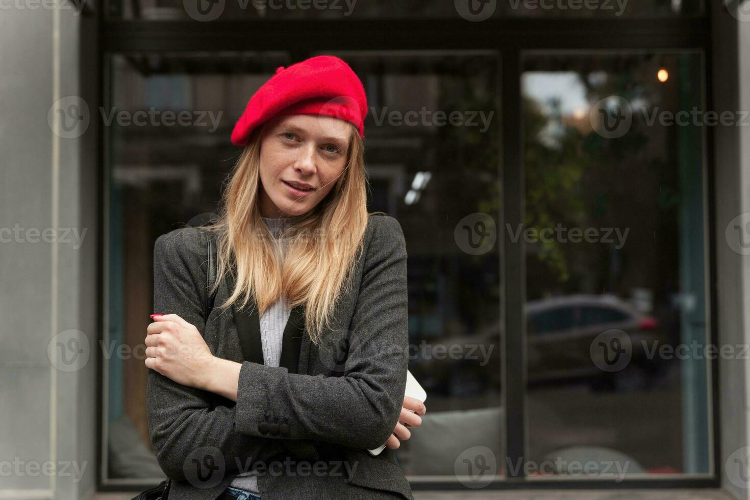 bellissimo giovane bionda lungo dai capelli femmina conservazione sua mani attraversato mentre in posa al di sopra di urbano sfondo nel elegante Abiti, conservazione smartphone nel sua mano foto