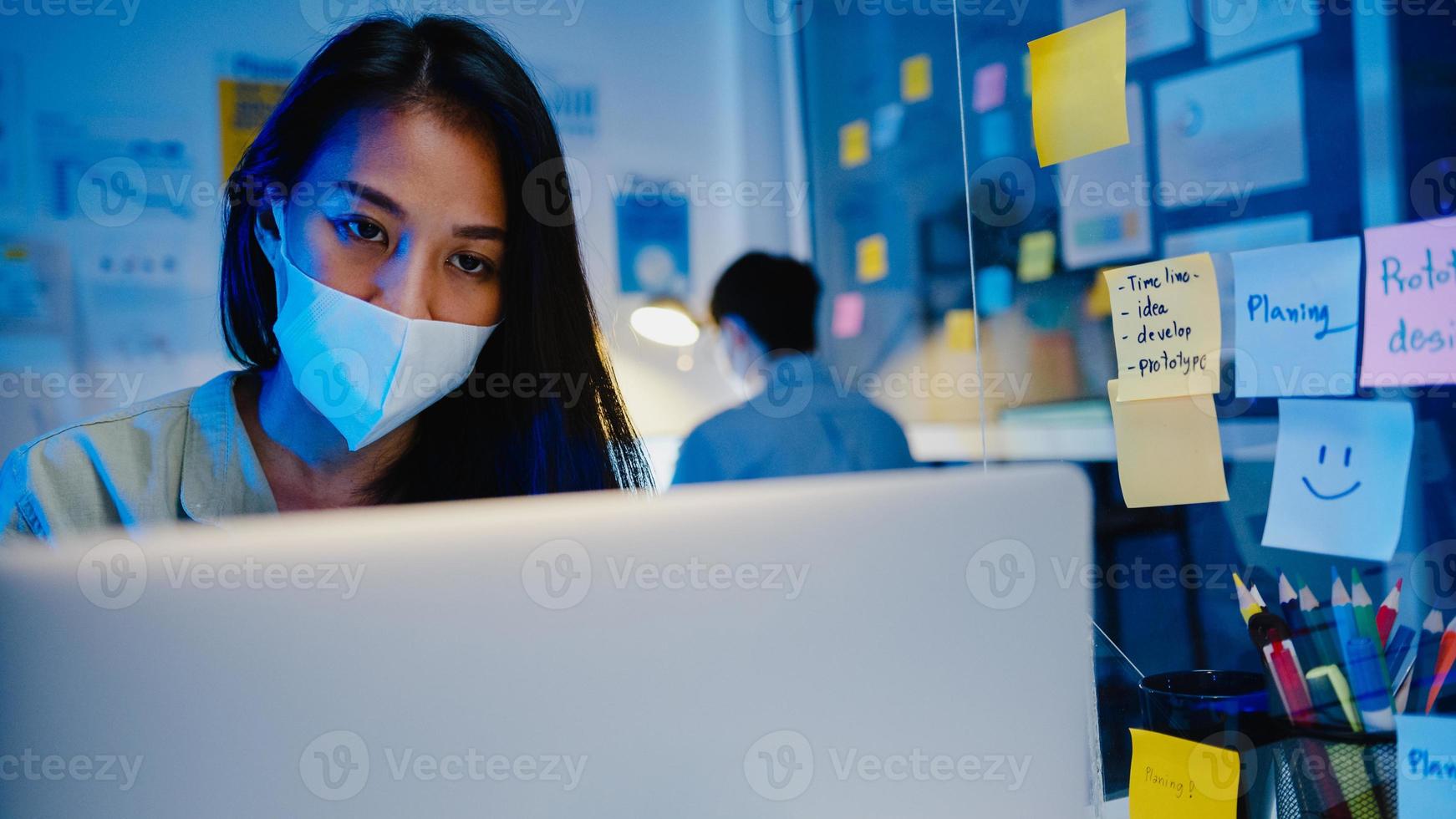 felice donna d'affari asiatica che indossa una maschera medica per il distanziamento sociale in una nuova situazione normale per la prevenzione dei virus mentre si utilizza il laptop al lavoro durante la notte in ufficio. vita e lavoro dopo il coronavirus. foto