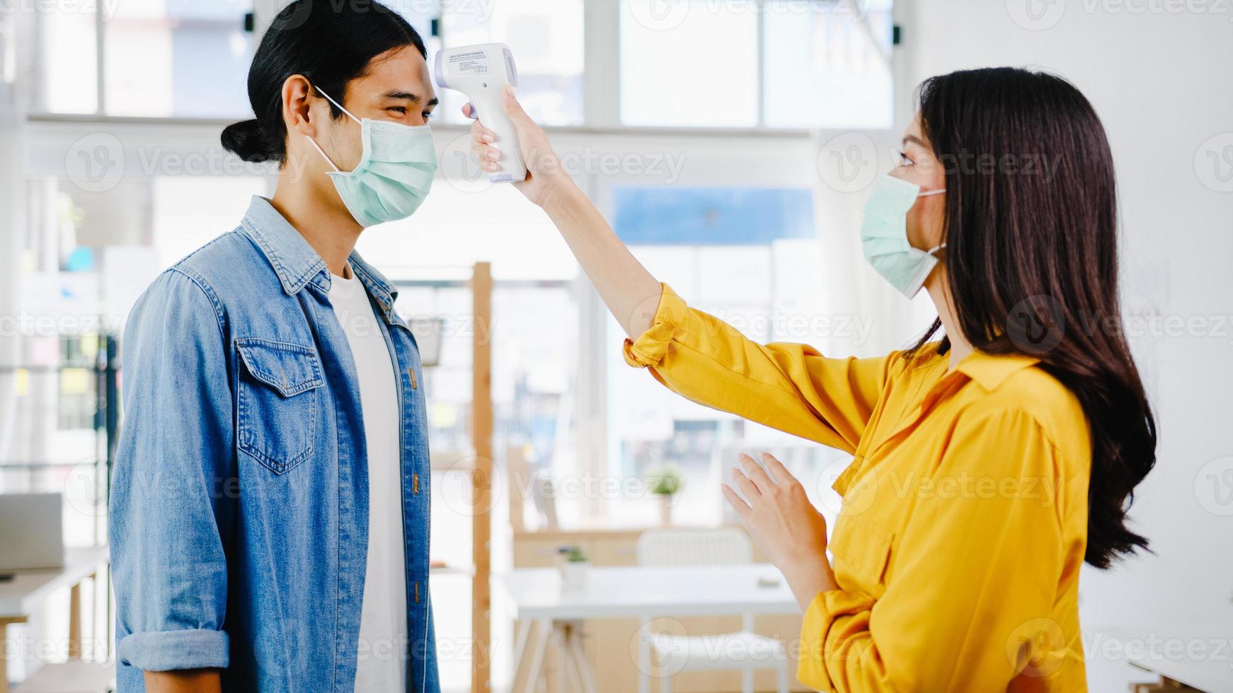 receptionist femminile asia che conduce indossare una maschera protettiva per il viso utilizzare un termometro a infrarossi o una pistola per la temperatura sulla fronte del cliente prima di entrare in ufficio. stile di vita nuovo normale dopo il virus corona. foto