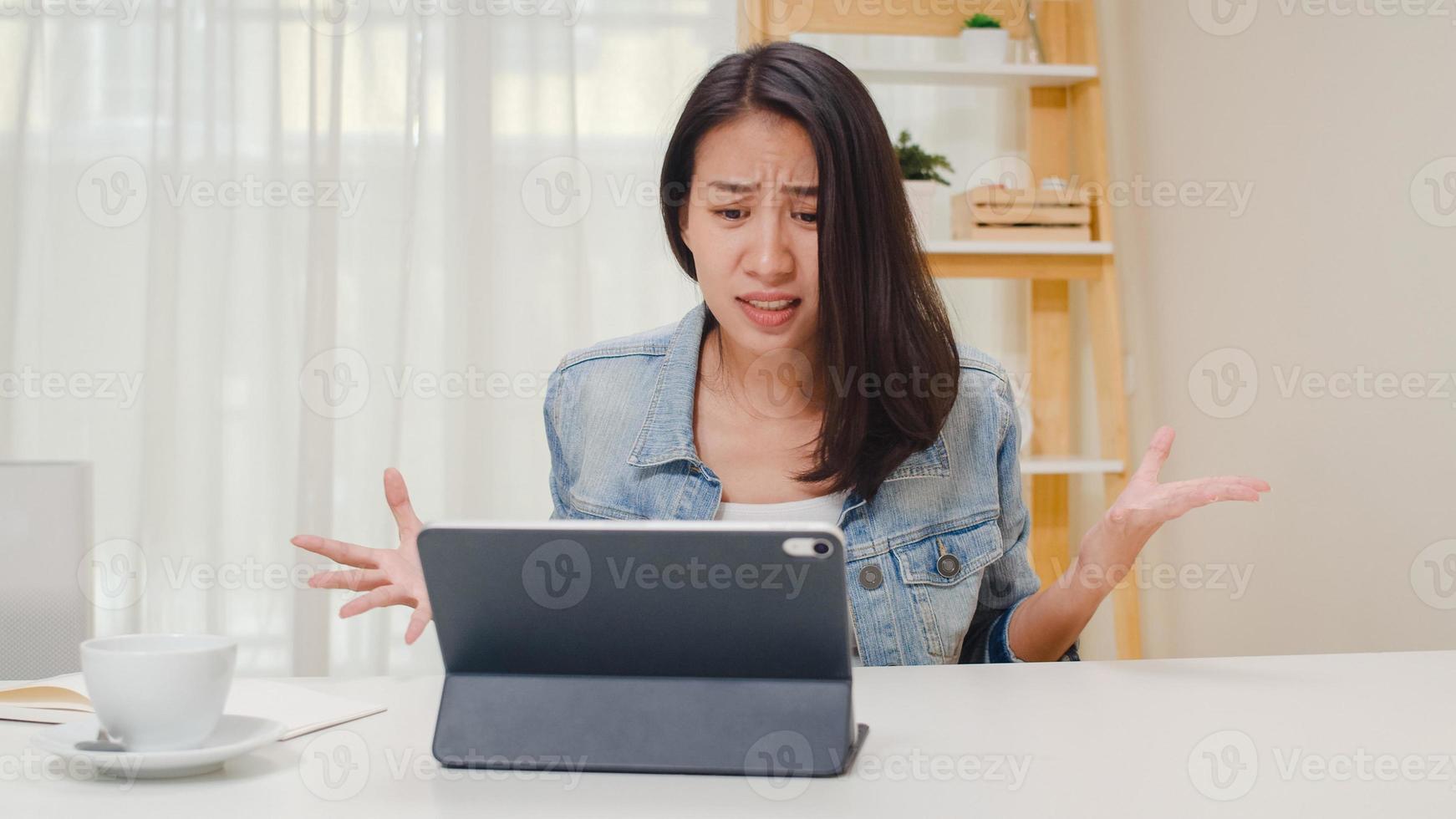 frustrata giovane donna asiatica che ha problemi con il computer tablet non funzionante seduto sulla scrivania. abbigliamento casual da donna d'affari intelligente freelance utilizzando tablet che lavora sul posto di lavoro in salotto a casa in ufficio. foto