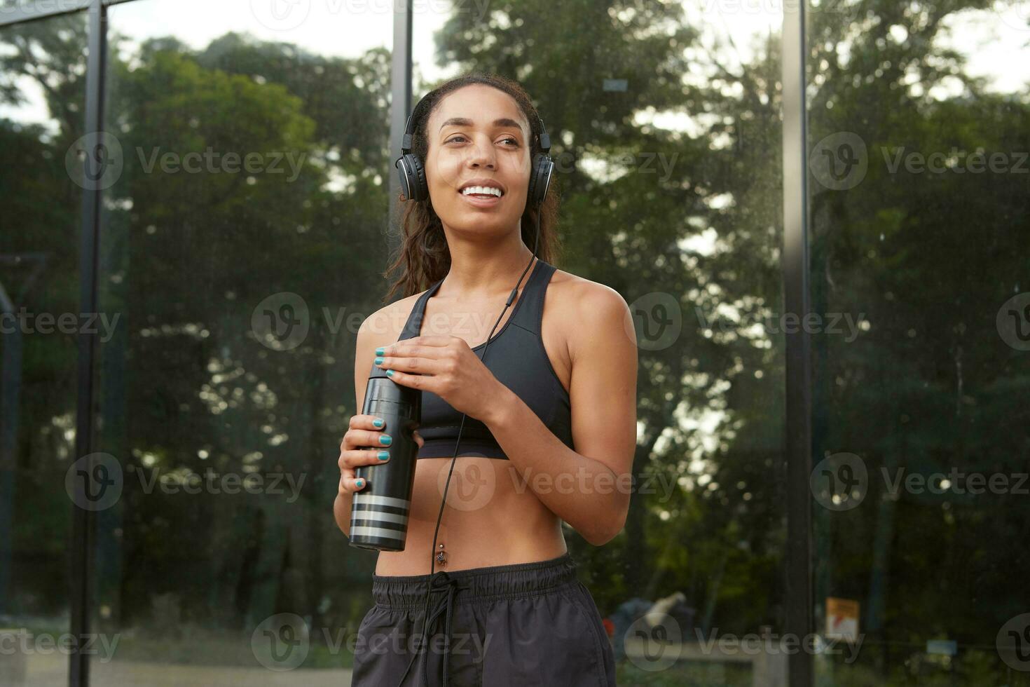 allegro giovane attivo buio spellato donna con coda di cavallo acconciatura indossare nero sportivo Abiti mentre fare yoga fitness esercizio nel strada, Tenere nero fitness bottiglia e sorridente felicemente foto