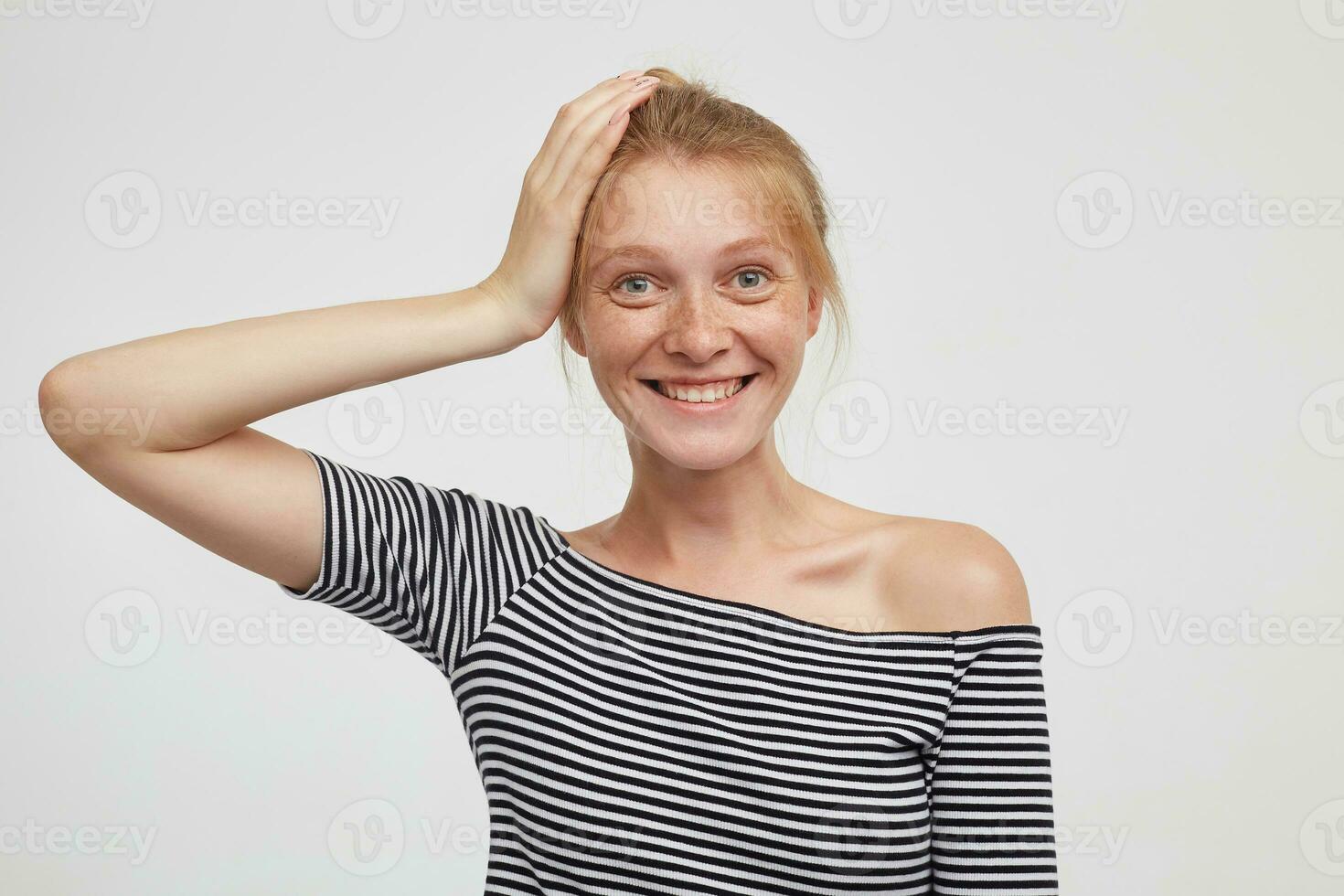 positivo giovane bellissimo testa Rossa femmina con naturale trucco conservazione sollevato palma su sua testa e guardare a telecamera con affascinante Sorridi, in piedi contro bianca sfondo foto