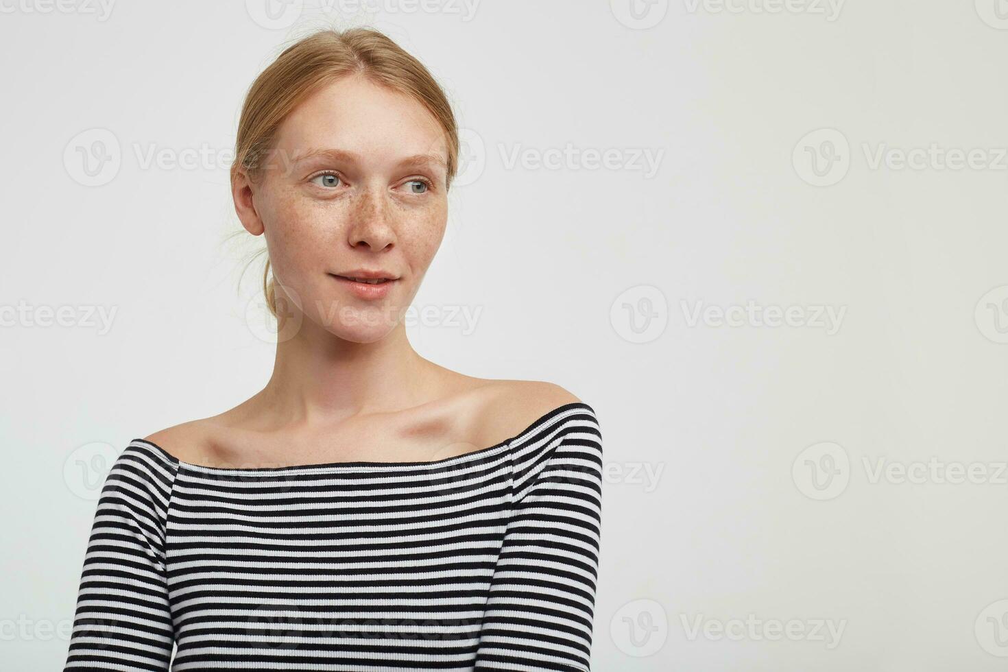 affascinante giovane bella testa Rossa signora con naturale trucco guardare pensieroso a parte con dolce Sorridi, vestito nel a strisce superiore mentre in piedi al di sopra di bianca sfondo foto