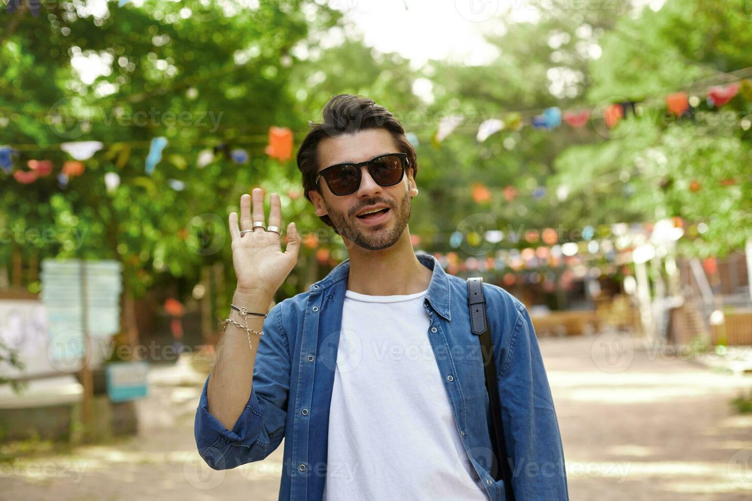 attraente giovane buio dai capelli uomo a piedi attraverso città giardino e raccolta mano nel benvenuto gesto, indossare casuale Abiti e occhiali da sole foto