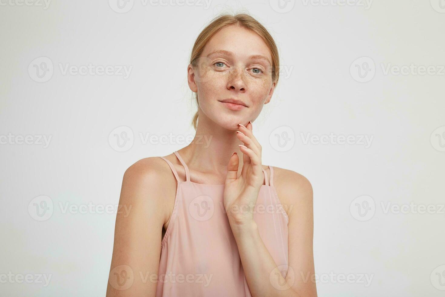 affascinante giovane bellissimo testa Rossa femmina guardare positivamente a telecamera con leggero Sorridi e toccante sua mento con sollevato mano, isolato al di sopra di bianca sfondo foto