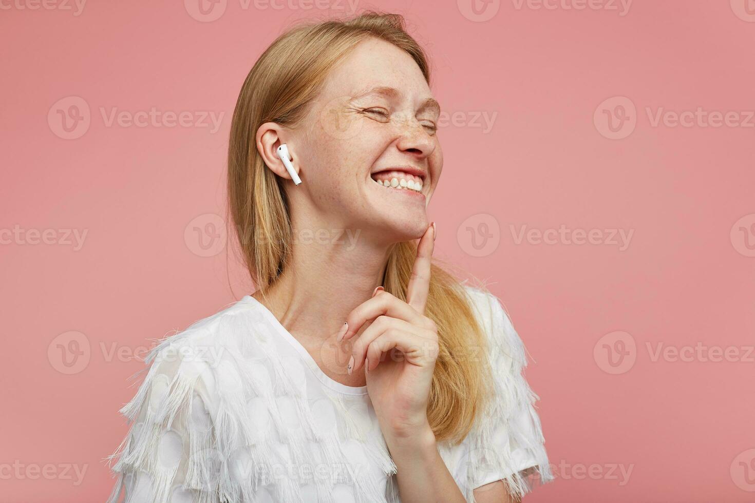 avvicinamento di piacevole guardare giovane bello testa Rossa donna con casuale acconciatura ridendo felicemente con chiuso occhi mentre ascoltando per musica nel sua auricolari, isolato al di sopra di rosa sfondo foto