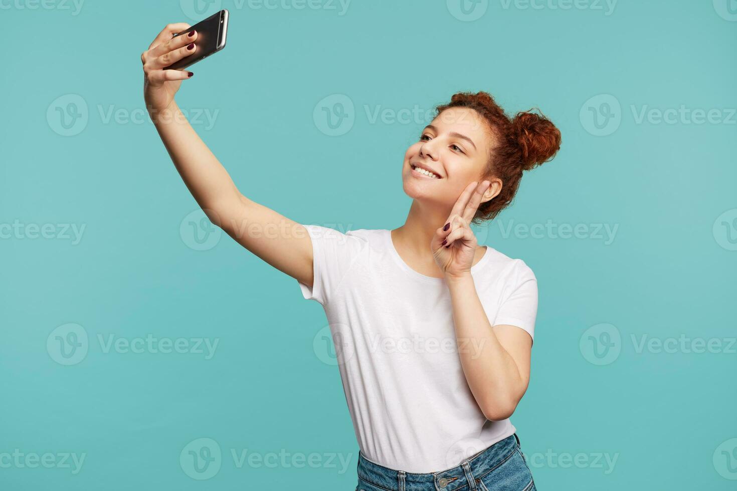 piacevole guardare giovane positivo Marrone dai capelli Riccio donna raccolta mano con vittoria gesto e sorridente allegramente mentre assunzione foto di se stessa su sua mobile Telefono, in posa al di sopra di blu sfondo