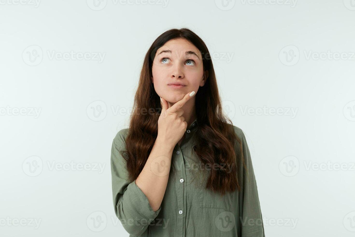 perplesso giovane bello lungo dai capelli signora con naturale trucco Tenere sollevato mano su sua mento mentre guardare pensieroso verso l'alto, essere ha riguardato di qualcosa mentre in posa al di sopra di bianca sfondo foto