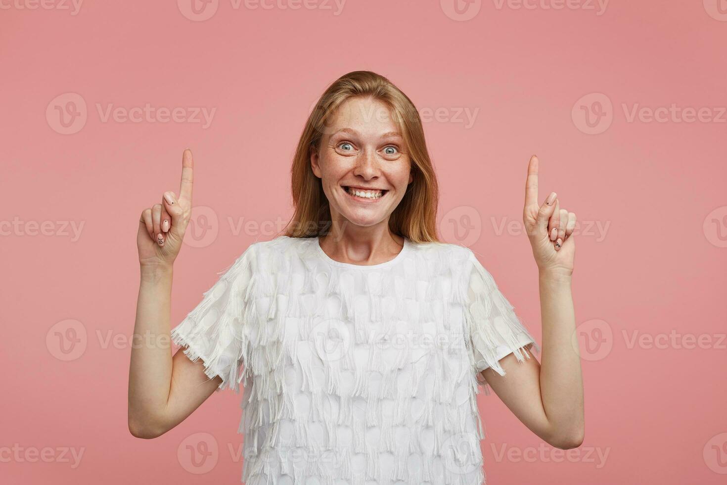 eccitato giovane bellissimo gioioso femmina con foxy capelli guardare a telecamera con agitato viso e sorridente in linea di massima, in piedi al di sopra di rosa sfondo con sollevato indice dita foto