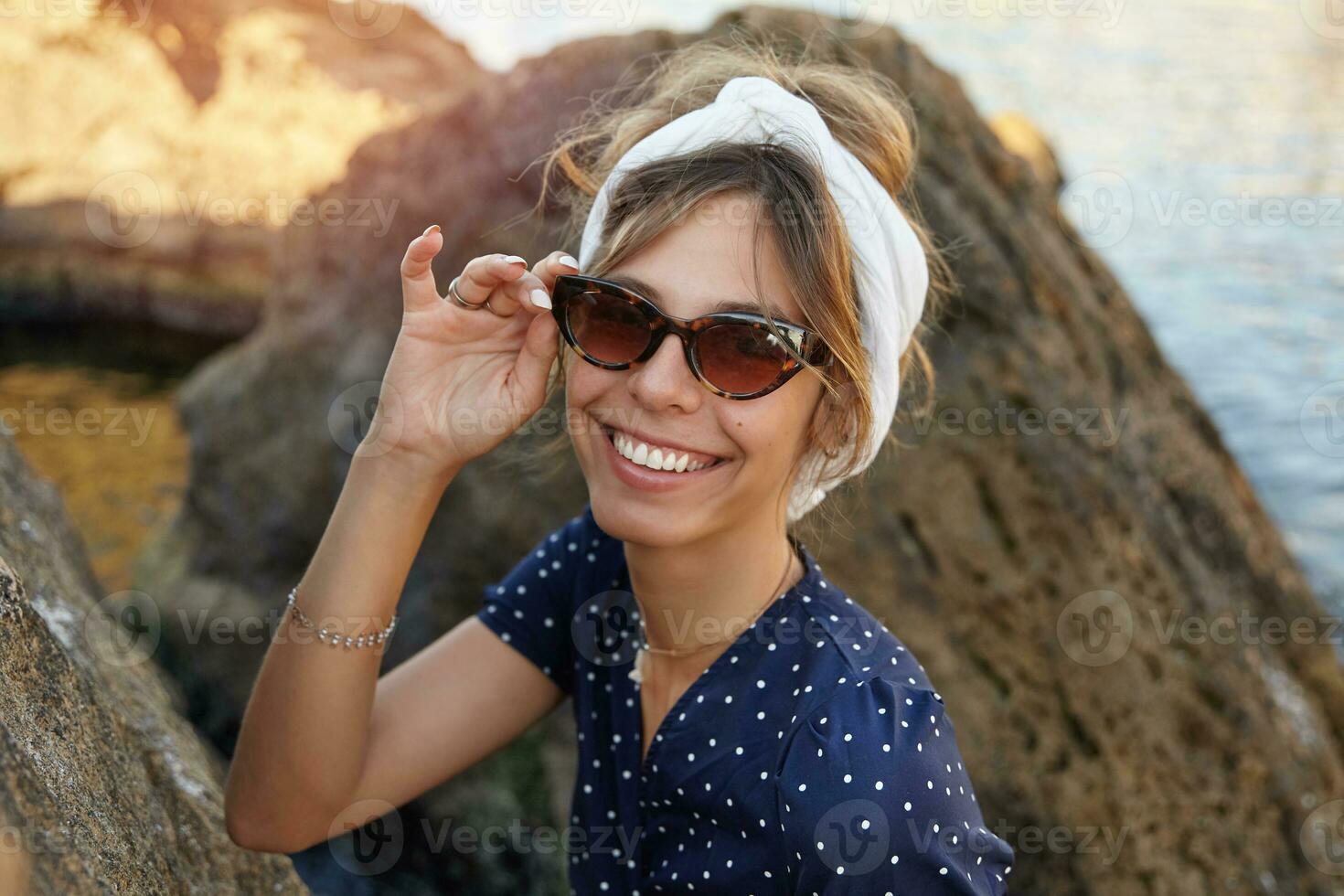 bellissimo giovane brunetta signora nel di moda occhiali da sole e bianca fascia per capelli in posa all'aperto su soleggiato giorno, sorridente con gioia per telecamera mentre seduta su grande pietra foto