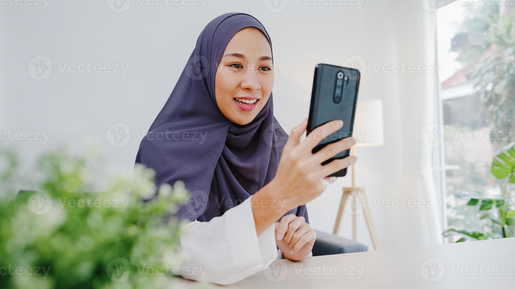giovane donna d'affari musulmana asiatica che utilizza lo smartphone parla con un amico tramite videochat brainstorming riunione online mentre lavora a distanza da casa in soggiorno. distanziamento sociale, quarantena per il virus corona. foto