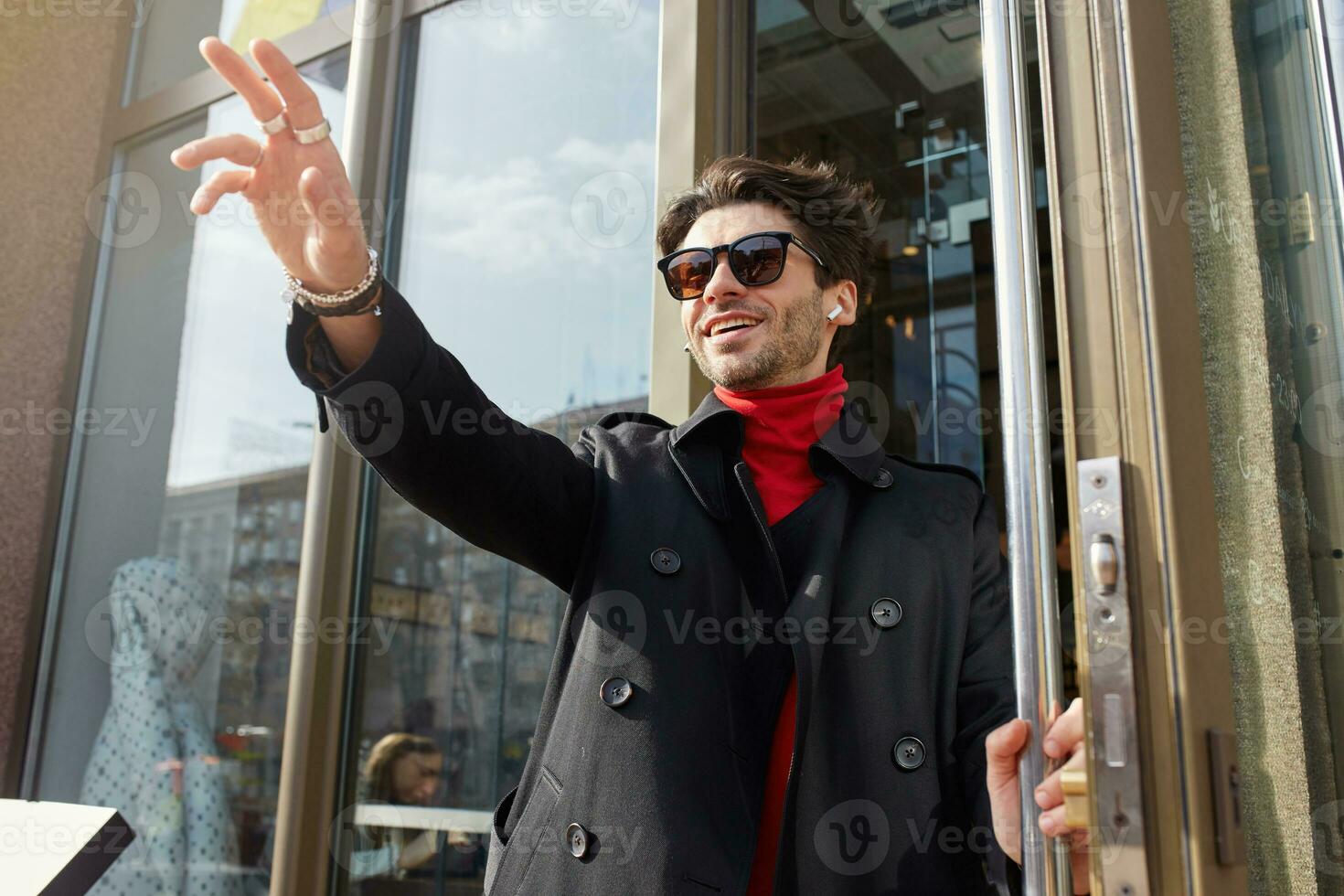 soleggiato tiro di giovane attraente Marrone dai capelli barbuto tipo nel di moda Abiti andando su di bar e incontro gli amici, raccolta mano nel Ciao gesto e sorridente felicemente foto