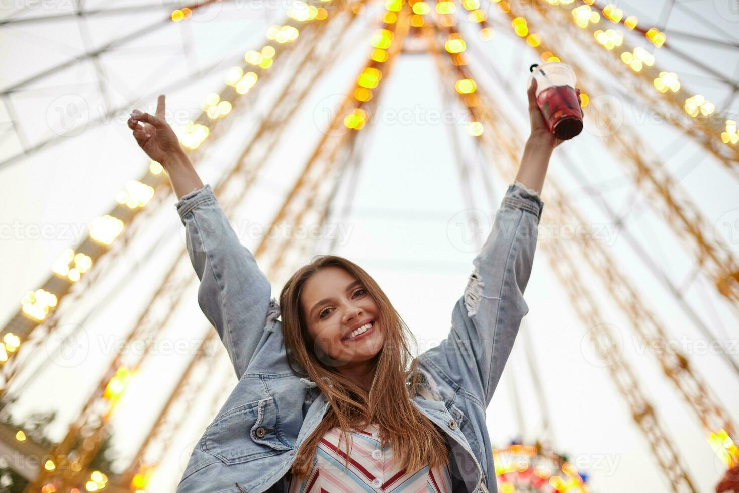 ritratto di bellissimo giovane donna con lungo Marrone capelli guardare per telecamera felicemente, in posa al di sopra di attrazioni nel città parco con sollevato mani, indossare casuale Abiti foto