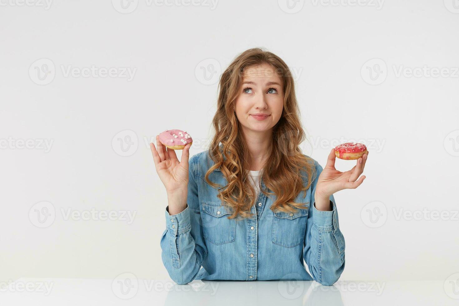 confuso giovane bella donna con lungo biondo ondulato capelli, seduta a il tavolo e detiene ciambelle nel sua mani. sembra su e meraviglie se per mangiare dolci per il notte. isolato al di sopra di bianca sfondo. foto