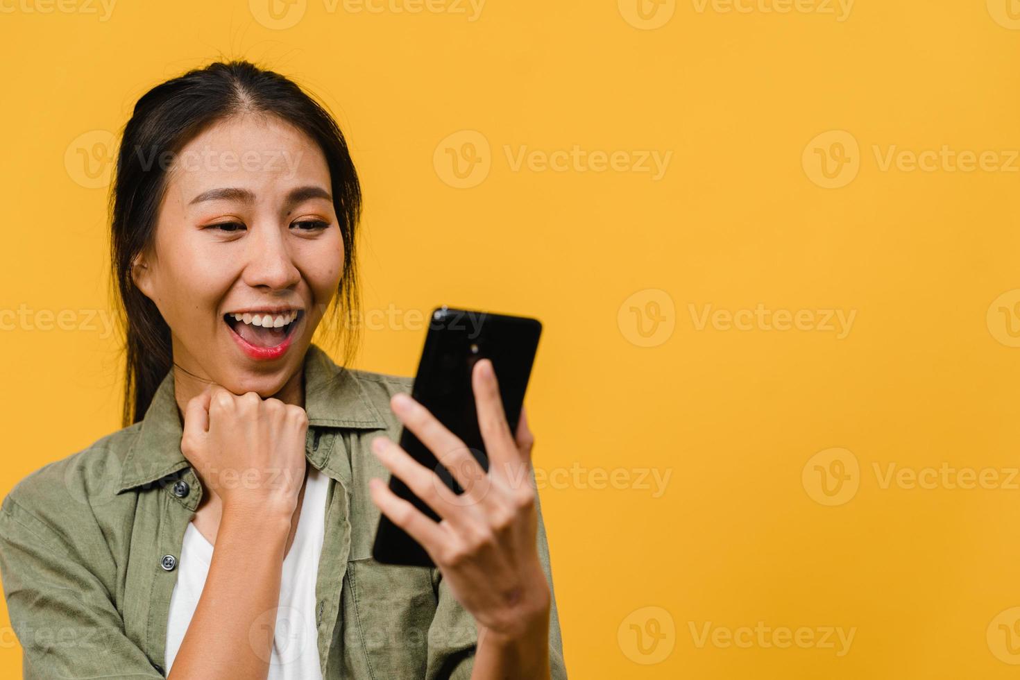 sorpresa giovane signora asiatica che utilizza il telefono cellulare con espressione positiva, sorride ampiamente, vestita con abiti casual e sta isolata su sfondo giallo. felice adorabile donna felice esulta successo. foto