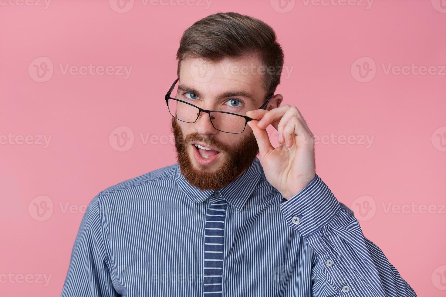 giovane allegro attraente dalla barba rossa giovane tipo con bicchieri e un' a strisce camicia, sembra a il telecamera attraverso bicchieri e sorrisi in linea di massima, sta al di sopra di rosa sfondo. foto