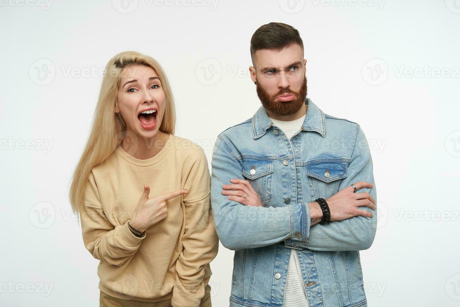 offeso giovane brunetta barbuto uomo pieghevole il suo mani su il petto mentre bella lungo dai capelli bionda femmina mostrando animatamente su lui con largo bocca ha aperto, isolato al di sopra di bianca sfondo foto