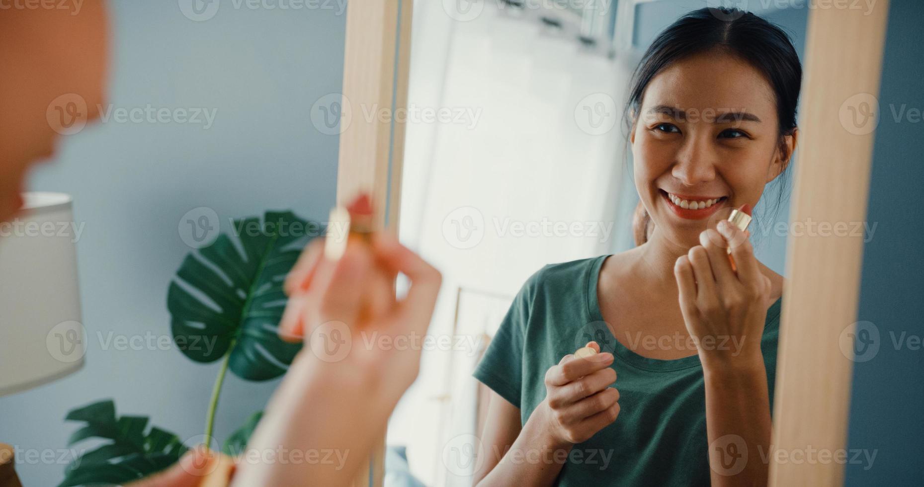 la bella signora asiatica con un panno casual si mette il rossetto sulle labbra davanti allo specchio in camera da letto a casa la mattina prima di uscire a uscire. giovane donna sorridente che applica trucco e che esamina specchio. foto