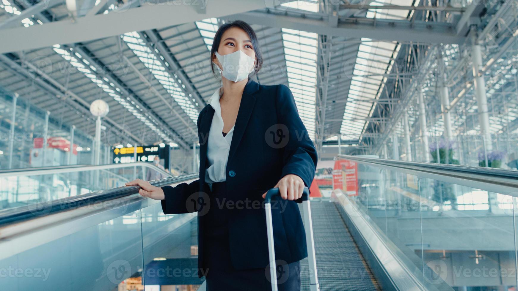 la ragazza d'affari asiatica indossa la maschera per il viso trascina il portabagagli sulla scala mobile guarda intorno a piedi fino al terminal dell'aeroporto internazionale. pandemia di covid per pendolari d'affari, concetto di distanza sociale di viaggio d'affari. foto