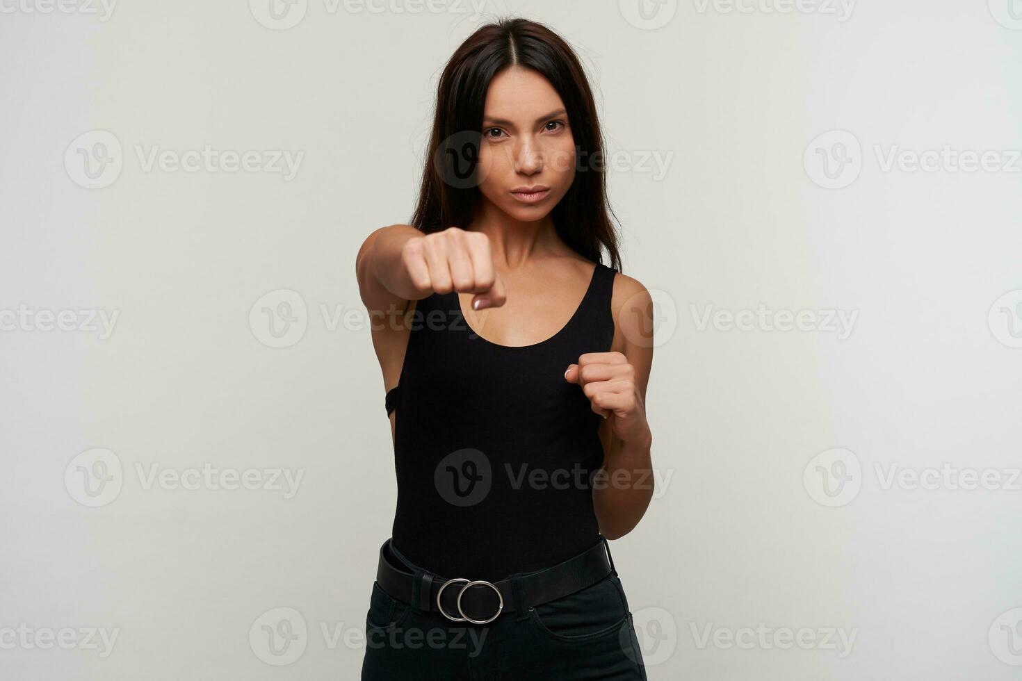 interno tiro di giovane bella buio dai capelli donna con casuale trucco boxe con sollevato pugni e guardare minacciosamente per telecamera, in piedi contro bianca sfondo foto