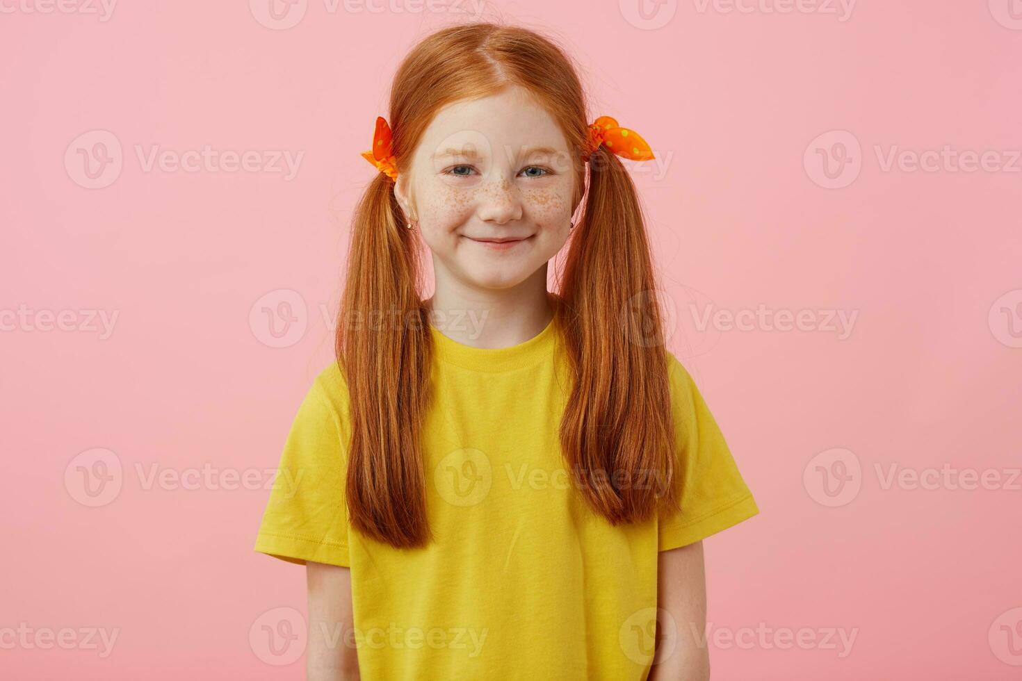 ritratto di petite lentiggini dai capelli rossi ragazza con Due code, sembra in il telecamera e sorrisi, indossa nel giallo maglietta, sta al di sopra di rosa sfondo. foto